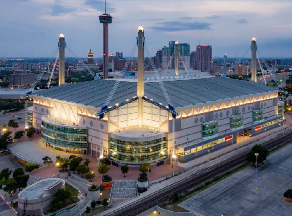 An aerial view of the Alamodome lit up against an evening grey sky with the San Antonio skyline behind it