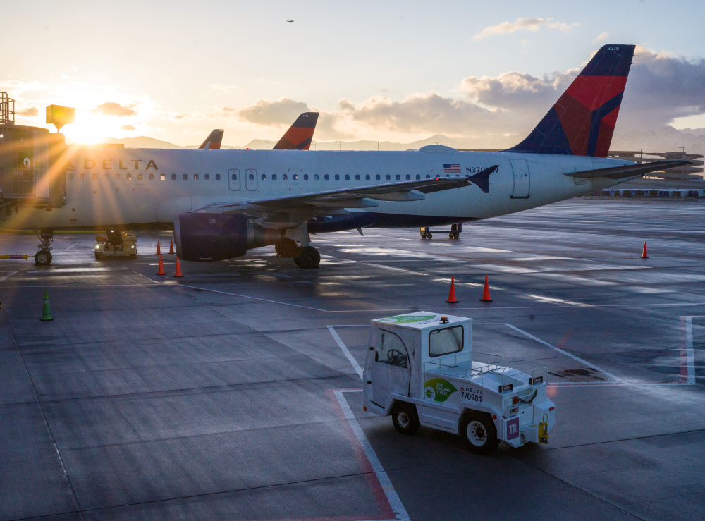 A Delta Airlines plane sits on the runway in front of a sunset sky with an electric baggage tractor heading towards it