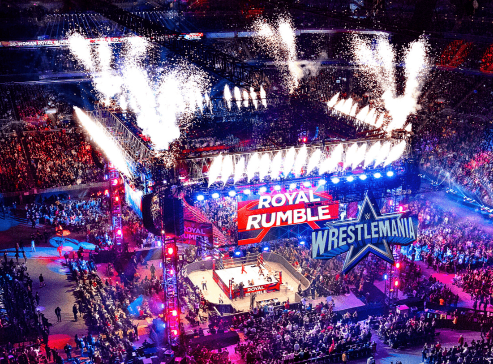 Royal Rumble WWE event in the center ring of a full stadium lit with blue and purple lights underneath white firework fountains