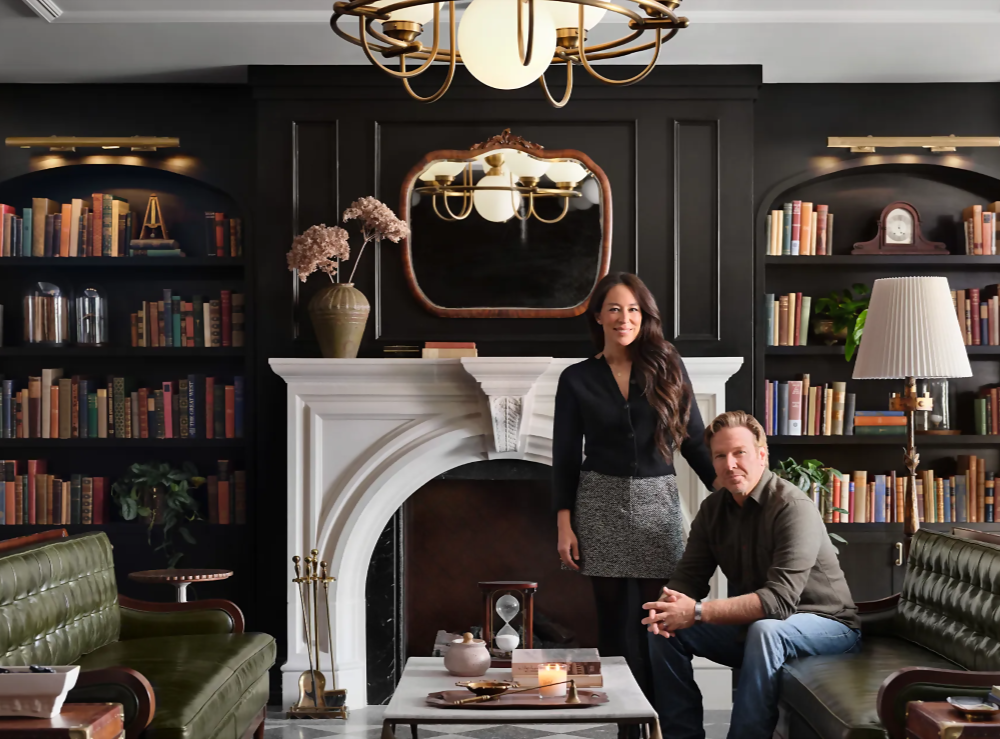A man sits on a sofa and a woman stands next to him in a library with classic style.